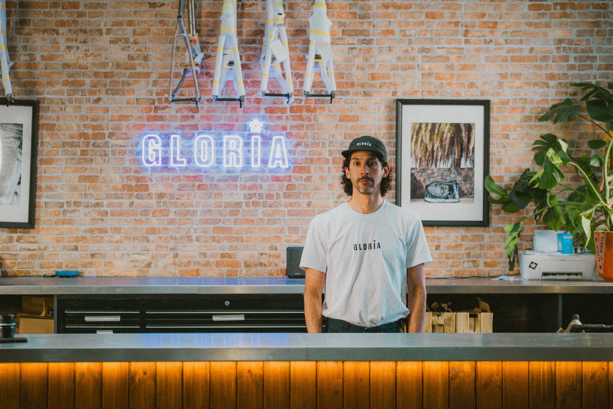 Person wearing a white t-shirt with 'Gloria' logo in front of a brick wall with neon sign and framed pictures.