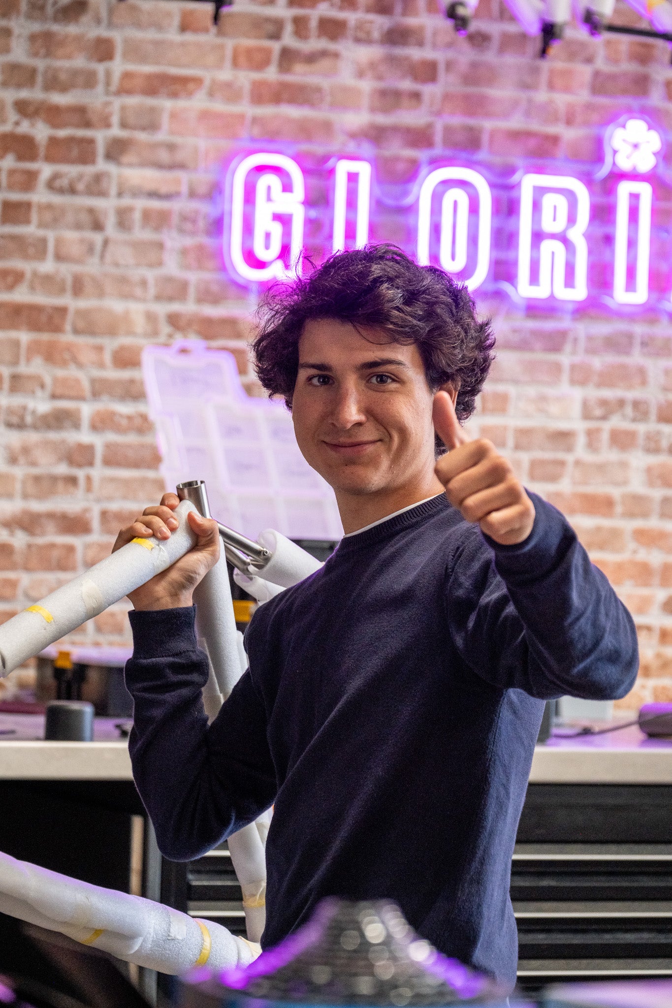 Person holding a bike in front of a brick wall with 'GLORIA' neon sign