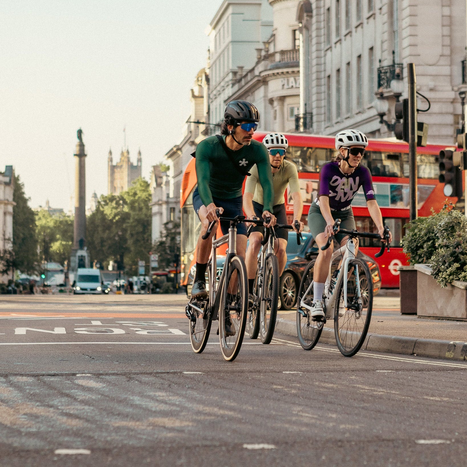 Three cyclists riding bicycles on a city street with a red double-decker bus in the background.